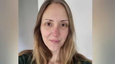 A woman with long blonde hair is looking at the camera. She has brown eyes and silver rings in her nose and lip. 