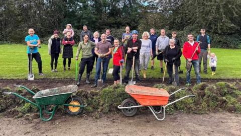 A large group of men and women carrying shovels stand on a patch of grass smiling at the camera. In front of them are two wheelbarrows .