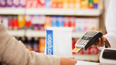 A generic getty image of someone paying for a prescription in a white bag at a pharmacy with medicines on shelves blurred in the background 