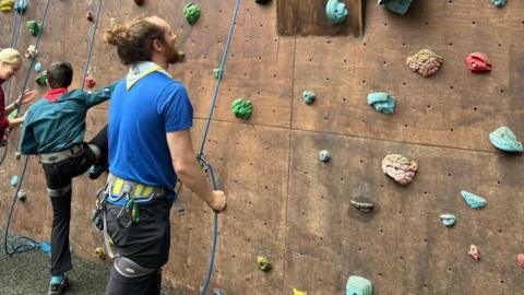 A man looking up at a climbing wall wearing a blue scouts top and neck scarf, with a young boy about to climb the wall next to him talking to a different man.