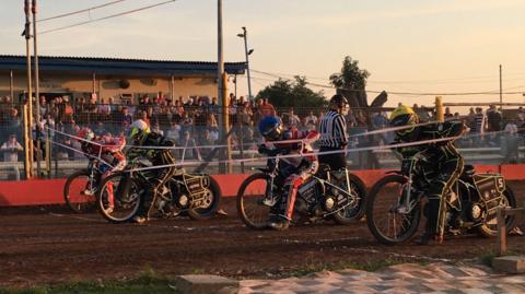 Riders are getting ready behind the starting line at Swindon Speedway. People are watching from the stands.