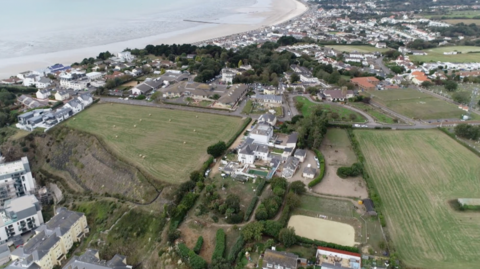 A sweeping aerial view shows the overdale site and winding roads. In the foreground, a patchwork of green and tan fields is divided by hedgerows and narrow lanes, some dotted with hay bales. A steep embankment or cliff edge separates part of the farmland from lower residential buildings. At the centre, a cluster of larger properties and outbuildings sits among trees, surrounded by smaller homes and winding roads. Further inland, the town becomes denser, with rows of houses and a street grid extending toward open countryside. The coastline arcs gently into the distance, giving a sense of the town’s layout between sea and rural land.