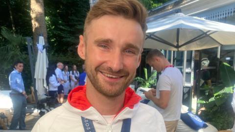 A man wearing a white jacket with red lining smiles for the camera with a medal around this neck while other people are stood in the background