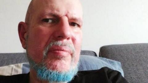 A bald man with a blue and grey coloured beard and wearing a black top, takes a selfie while sitting on a grey sofa, with a blue and white cushion behind him.