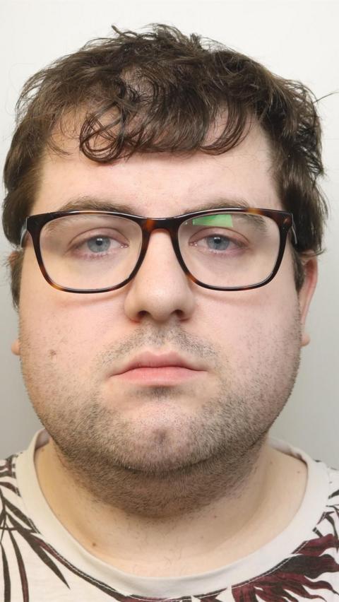 Bennett has short dark hair and is wearing glasses. He has stubble and a t-shirt on. It is a mugshot of his face taken while in police custody