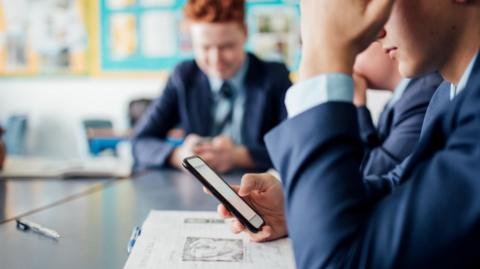 Young male student in a blue school uniform is pictured checking his phone in a classroom.