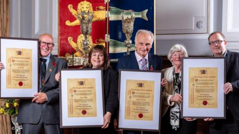 Recipients of the Freedom of the Borough stand from left - Cecil Page of the Kingfisher Boxing Club, Jayne Biggs of Norfolk Heart2Heart, Hugh Sturzaker of the Great Yarmouth Civic Society and members of the Great Yarmouth branch of The Samaritans. They are holding their scrolls of honour, mounted in frames, and stand before the staff and sceptre and the borough crest of Great Yarmouth. It is a shield painted red and blue divided vertically, with lion heads over the red segment, and herring fish tails over the blue, the heraldic image granted by King John in 1208.