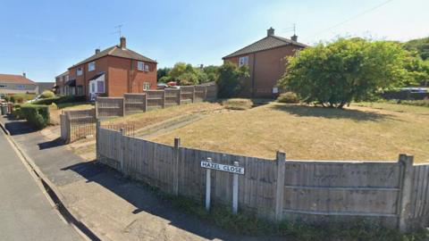 Hazel Close is a residential road with hedges and gardens and there are cars parked in driveways. The road sign is near a raised, fenced lawn, which has a tree in the middle of it.