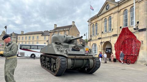 A military green Sherman tank sits outside the town hall in Melksham, a portion of the hall has been decorated with a cascade of poppies.