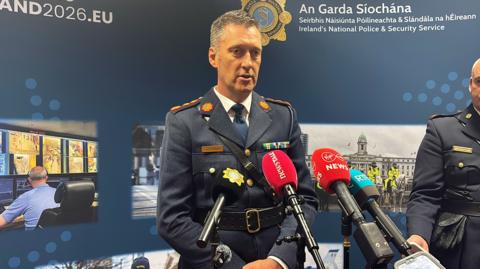 Garda officer wearing a blue uniform. He has short dark and grey hair and has mics in front of him. An Garda Siochana is on the background behind with police pictures.
