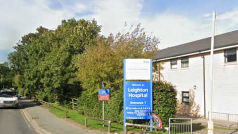 A blue NHS sign that reads Leighton Hospital, with a road running to left od the sign and a white building on the right. A green hedge is in the background next to the road. 
