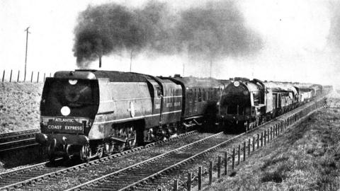 A black and white picture of the locomotive in 1950, it has smoke coming out of the top as it travels down a railway track with grass hills either side, there is a sign at the front of the locomotive which says Atlantic Coast Express