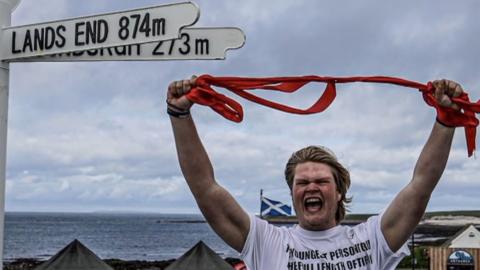 Marcus, aka The Hull Boy, with an expression of joy while holding up a red ribbon, standing by a sign saying 'Land's End 874m'