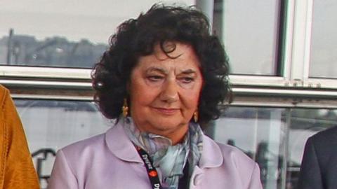 Caroline Jones, a woman with dark curly hair wearing a pink jacket.