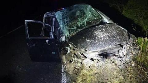 The badly damaged car. The front section is crushed, while the bonnet is heavily scratched and the windscreen smashed. The right-hand front door is open and again the glass is smashed.