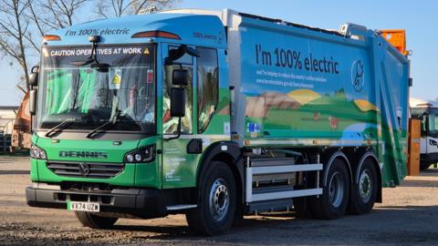 A waste collection lorry rests on gravel. Emblazoned on the side and front are the words "I'm 100% electric".