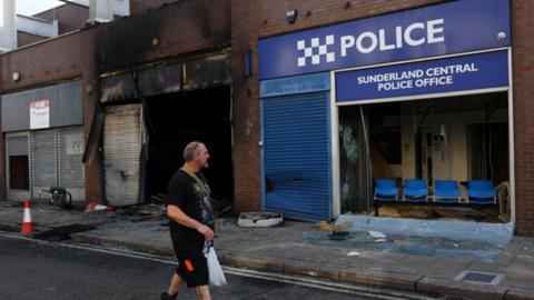 Burned-out building next to blue police building saying "Sunderland Central Police Office".