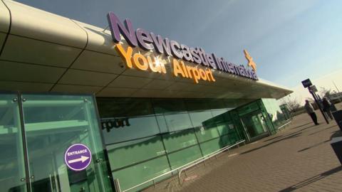 Newcastle Airport. A large glass building with a sign on the front of the roof reading 'Newcastle International Your Airport' in orange and purple lettering. There is a blue sky and two people in the distance standing on the pavement. 