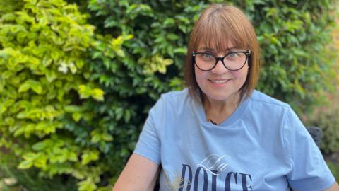 Catherine Turnbull is smiling at the camera. She is wearing a pale blue t-shirt and dark framed glasses. Catherine has short light brown hair. She has some bushes behind her with are slightly out of focus. 