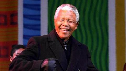 Nelson Mandela grins as he dances on stage at a concert in Leeds in 2001. There are singers behind him with a backdrop of green, blue and orange colours celebrating African culture.