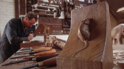 A man sits in a woodworking workshop. In the forefront is an oak bookend with a mouse carved into it.