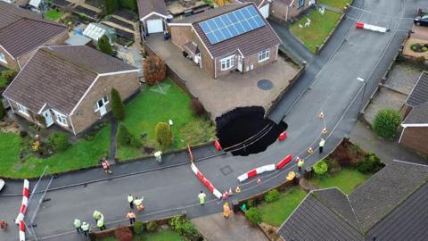 Aerial shot of large sinkhole in street on the drive of one house and into the road, with other homes nearby, barriers in the street, and workers standing around