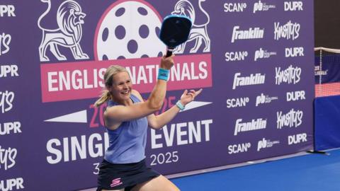 Thaddea is in a blue vest and blue skort, with her racket in the air, backed by a purple sponsorship board.