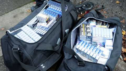 Two black cross body bags containing packets of illegal cigarettes, tobacco and vapes which have been seized by police from shops in Northampton. Both bags are full to the brim of items.