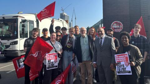 Former Labour leader Jeremy Corbyn waring a light coloured shirt, brown trousers and dark jacket. He is next to a man in a suit and they are surrounded by a group of protesters, some who are holding posters and red flags. 