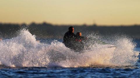 A water scooter with two people visible on board throws up white spray on a open stretch of water