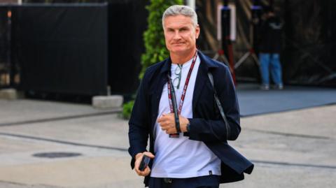 A man with short grey hair walks outdoors in a paved area, wearing a white T-shirt under a dark jacket and trousers. He has a lanyard around his neck, holds a phone in one hand, and has glasses hanging from his shirt, with equipment and greenery visible in the background.