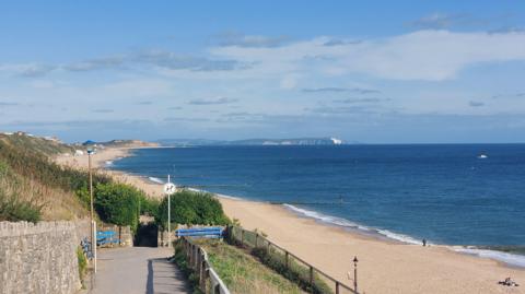 A long stretch of golden beach, lapped by sea waves, can be seen from the cliff above stretching towards the horizon, with blue skies above