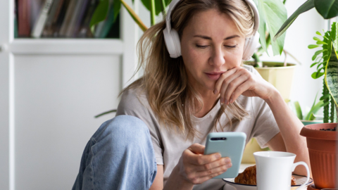 A stock shot of a young blonde woman wearing white headphone and looking at her phone.