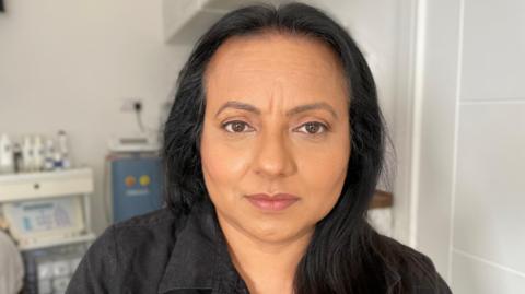 Sukhjeet Bahra looks directly at the camera. She has dark black hair and is wearing a black shirt with a collar. 