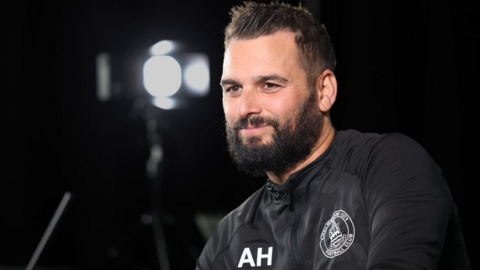 Angelo Harrop, wearing a Chelmsford City black tracksuit top, sitting for down with an interview with lighting in the background.