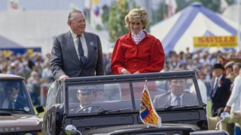 A man and a woman stand in the back of an open top car as they move through a rural show. The man wears a grey suit while the woman is wearing a red jacket with a red and white polka dot blouse. 