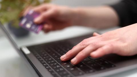 An out of focus stock image of a person typing on a laptop with one hand, and holding a credit card in their other hand.