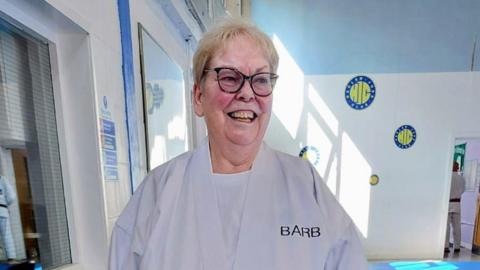 Barbara Carolan has short fair hair and is wearing glasses. She is wearing a judo suit, standing in a judo hall, and looking just off-camera, laughing.
