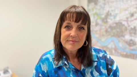 A woman with shoulder length dark hair and a fringe smiling at the camera. She is wearing a blue blouse and there is an out of focus map behind her.