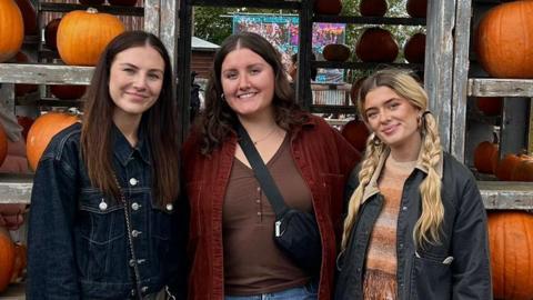 Three girls in their early 20s smile with pumpkins behind them on shelves.