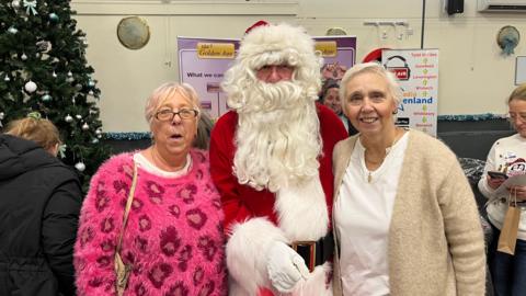 Jane Van Der Merwe, left, with her sister Gail Lewis, and Santa Claus in the middle. Jane has short blonde hair with a pink tint and is wearing oval-shaped glasses and a pink furry jumper with red patterns on it. Gail has short grey hair and is wearing a white top with a beige cardigan on top. There is a Christmas tree behind them on the left.