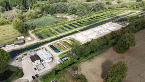 A drone shot of the site, with a series of ponds surrounded with greenery going down one side of a large channel which lookts like a river but isn't. On the other side, a couple of smaller industrial buildings and around six concrete tanks.