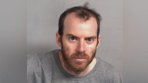 A custody shot of a man looking directly at the camera while wearing a grey jumper. He has a brown beard and hair. 