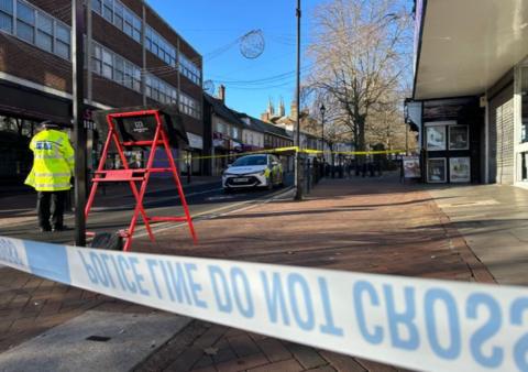 A close up of the reverse of some police tape at the end of a high street with a police car and a police officer in a high-vis jacket in view in the distance