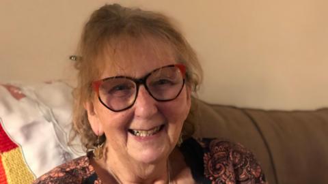 Judy Arliss is smiling, her light brown hair is tied back and she is wearing black and red glasses. She has dangly earrings in and a red paisley patterned top.