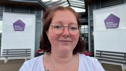 Heather Cliffe is outside a building at Bishop Grosseteste University. She has round gold glasses and plum coloured hair shaped into a bob.
