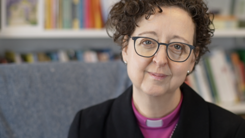 The Bishop of St Edmundsbury and Ipswich, Dr Joanne Grenfell, smiles at the camera as she sits in a room with bookshelves behind her. She has short, dark curly hair and wears a black coat with a pink clerical top underneath and a white clerical collar. She is also wearing glasses.
