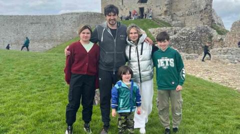 The family are standing in a row in leisure wear in front of the ruins of a castle. They are smiling for the camera and dad Alex has his arms around his wife and two oldest children while the youngest stands in front of them.