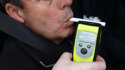 A man who is sat in the drivers seat of a car with his seat belt on. He is undertaking a breathalyser test.