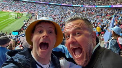 Two men smiling toward the camera, behind them are football fans. They both have their mouths wide in an excited look.